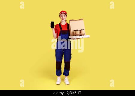 Une femme de messagerie tient le café et les boîtes avec pizza, offrant des boissons et de la livraison de nourriture, montrant écran de téléphone mobile vierge pour la publicité. Studio d'intérieur isolé sur fond jaune Banque D'Images