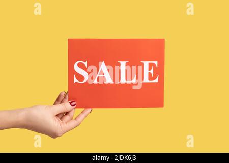 Profil vue de côté gros plan de la main de femme tenant la carte de vente rouge, méga shopping, réductions pour le vendredi noir. Studio d'intérieur isolé sur fond jaune. Banque D'Images