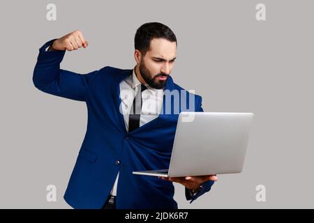 Homme puntant l'écran d'un ordinateur portable, regardant avec furieuse expression folle, boxe menaçant de frapper tout en ayant la conversation en ligne, portant le costume de style officiel. Prise de vue en studio isolée sur fond gris Banque D'Images