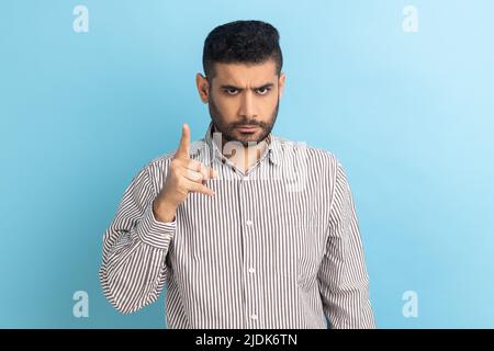 Portrait d'un homme d'affaires barbu sérieux et strict debout avec un geste d'admonage, en train de se grimader pour erreur et avertissement, portant une chemise rayée. Studio d'intérieur isolé sur fond bleu. Banque D'Images