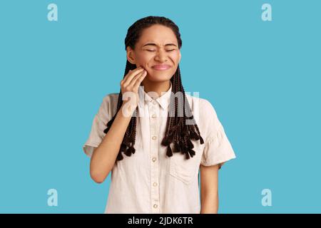 Une femme mécontentée souffre de maux de dents, touche la joue, a besoin de voir le dentiste, a des problèmes de santé, ferme les yeux avec la douleur, porte une chemise blanche. Studio d'intérieur isolé sur fond bleu. Banque D'Images