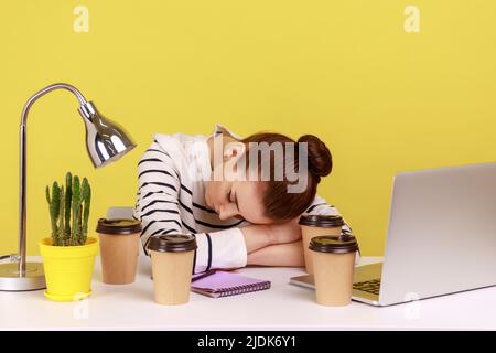 Femme fatiguée, directrice de bureau dormant sur le lieu de travail, couchée au milieu de tasses de café, se reposant après des heures supplémentaires. Studio d'intérieur tourné isolé sur fond jaune. Banque D'Images