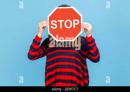 Femme inconnue portant un chandail rayé de style décontracté, couvrant le visage avec le symbole Stop, tenant le signe rouge, avertissement de danger, restriction et limites. Studio d'intérieur isolé sur fond bleu. Banque D'Images