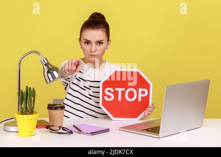 Femme sérieuse responsable de bureau portant une chemise rayée sur le lieu de travail devant l'ordinateur portable, tenant le panneau d'arrêt rouge et pointant le doigt vers l'appareil photo. Studio d'intérieur tourné isolé sur fond jaune Banque D'Images