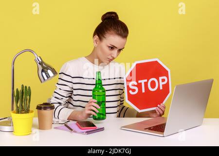 Femme employée de bureau dans une chemise rayée, assise sur le lieu de travail et ayant une vidéoconférence, montrant une bouteille d'alcool et un panneau d'arrêt rouge, studio d'intérieur tourné isolé sur fond jaune. Banque D'Images