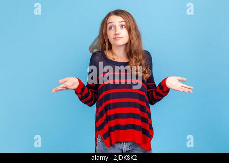 Je ne sais pas. Confus jeune belle femme portant rayé style décontracté chandail haussant les épaules, confus et incertain avec l'information. Studio d'intérieur isolé sur fond bleu. Banque D'Images