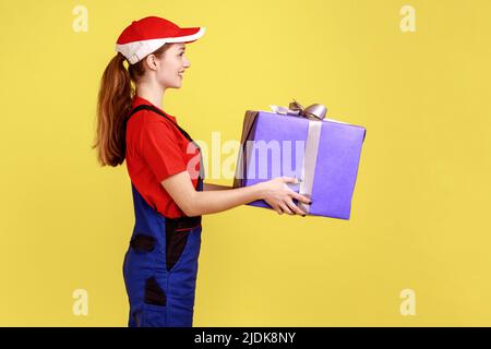 Vue latérale d'une femme à livraison positive debout tenant la boîte à cadeaux, livrant des cadeaux pour les vacances, portant une combinaison et une casquette rouge. Studio d'intérieur isolé sur fond jaune. Banque D'Images