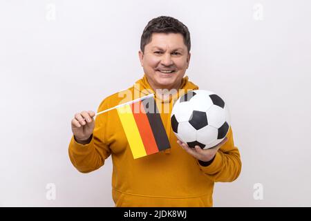 Homme d'âge moyen avec un sourire crasseux tenant le drapeau de l'allemagne et le ballon de football noir et blanc classique et regarder le match, portant le sweat à capuche de style urbain. Studio d'intérieur isolé sur fond blanc. Banque D'Images
