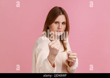 Portrait d'une femme blonde en colère, confiante et attirante, qui claque des poings, montrant un geste de boxe et prête à punch, portant un chandail blanc. Studio d'intérieur isolé sur fond rose. Banque D'Images