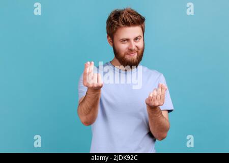 Portrait de l'homme barbu mercantile en train de frotter les doigts montrant le geste d'argent, demandant le salaire, exigeant des pots-de-vin, regardant la caméra avec le sourire. Studio d'intérieur isolé sur fond bleu. Banque D'Images