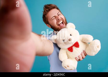 Portrait de l'homme barbu blogueur faisant selfie ou diffusant en direct, point de vue photo, tenant doux ours en peluche et rire. Studio d'intérieur isolé sur fond bleu. Banque D'Images