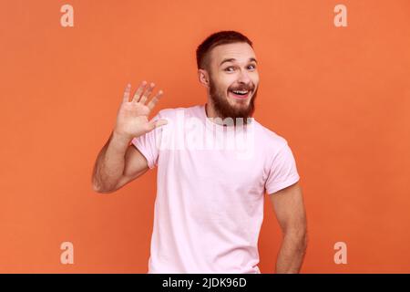 Portrait d'un bel homme barbu positif agitant la main et souriant amical à l'appareil photo, faisant un geste de bienvenue élevé, portant un T-shirt rose. Studio d'intérieur isolé sur fond orange. Banque D'Images