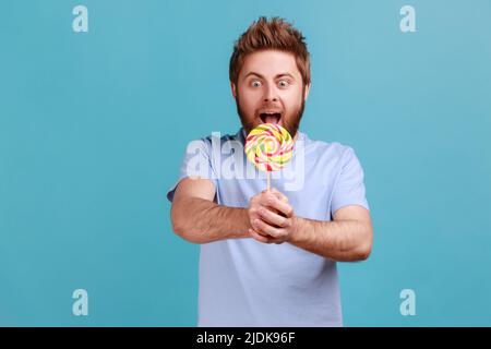 Portrait de l'homme barbu, impressionné et affamé, tenant des sucreries sucrées, se sent affamé, appréciant la délicieuse sucette. Studio d'intérieur isolé sur fond bleu. Banque D'Images