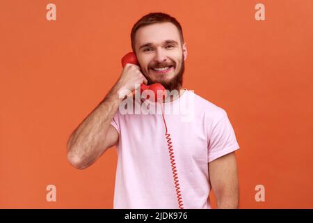 Portrait d'un homme barbu parlant téléphone fixe tenant dans le combiné, regardant l'appareil photo avec un sourire crasseux, portant un T-shirt rose. Studio d'intérieur isolé sur fond orange. Banque D'Images