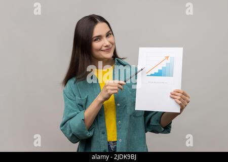 Portrait d'une femme d'affaires positive tenant un graphique en papier montrant l'augmentation des salaires des travailleurs, pointant vers le papier, portant une veste de style décontracté. Prise de vue en studio isolée sur fond gris. Banque D'Images