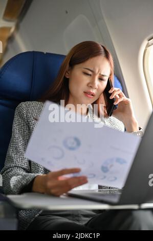 A souligné la femme d'affaires asiatique du millénaire, parlant au téléphone avec sa secrétaire, vérifiant son rapport d'affaires, travaillant à distance pendant le vol. Banque D'Images