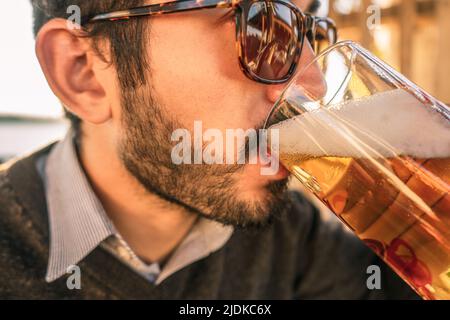 Jeune homme avec une barbe et des lunettes de soleil buvant de la bière dans une tasse Banque D'Images