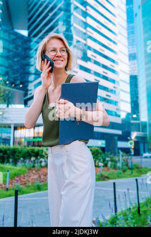 Portrait d'une femme d'âge moyen souriante avec des cheveux justes debout en centre-ville, tenant un ordinateur portable, parlant sur un smartphone. Banque D'Images