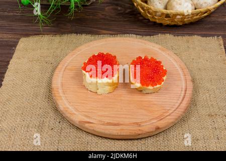 Délicieux sandwiches au caviar rouge et au beurre sur une planche de bois sur fond d'herbes et de champignons. Banque D'Images