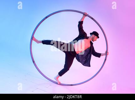 Corps complet d'acrobate masculin sportif en chapeau exécutant le stunt de valse avec roue cyr pendant l'entraînement sur fond clair en studio Banque D'Images