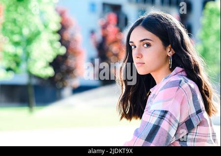 Une adolescente calme avec des cheveux noirs regardant l'appareil photo tout en étant assise dans la rue avec des arbres sur un arrière-plan flou dans la ville