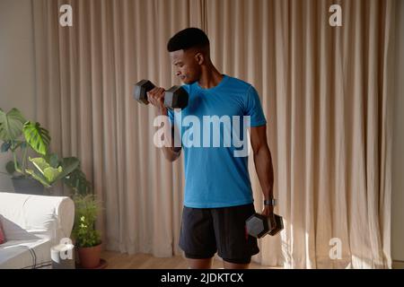 Un jeune homme noir afro-américain s'est fait travailler dans le salon à la maison en faisant une boucle de biceps avec de lourds poids de haltères. Maintien de la forme et de la santé avec Banque D'Images