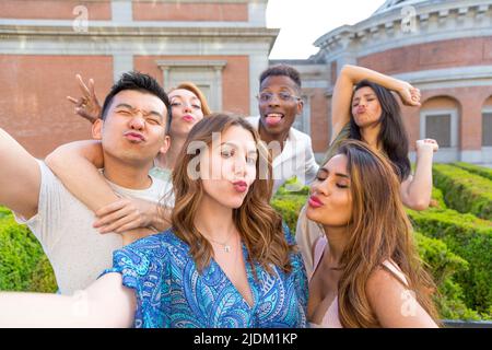 Groupe d'amis multiethniques posant soufflant un baiser d'une manière drôle tout en prenant un selfie à l'extérieur Banque D'Images