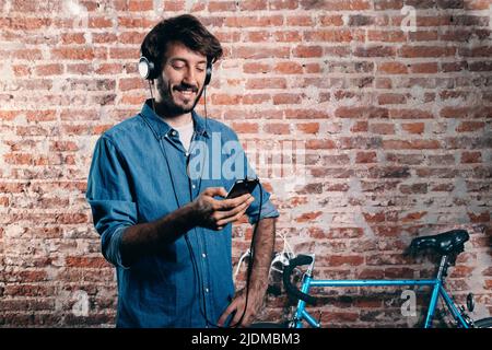 Jeune millénaire Écoutez de la musique avec un téléphone portable et un casque. En arrière-plan, un vélo bleu appuyé contre le mur de briques. Banque D'Images
