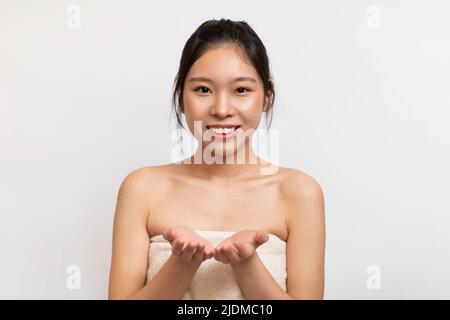 Bonne jeune femme asiatique enveloppée dans une serviette de bain démontrant quelque chose sur des palmiers vides, montrant un objet invisible Banque D'Images