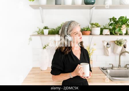 Femme écoutant attentivement les écouteurs tenant une tasse dans la cuisine avec des plantes Banque D'Images