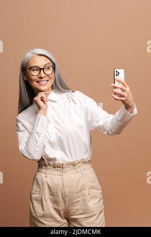 Une femme âgée heureuse utilise un smartphone pour la connexion vidéo isolée sur beige, insouciante mature grise-cheveux dame a satisfait la réunion virtuelle, grand-mère moderne faisant le selfie Banque D'Images