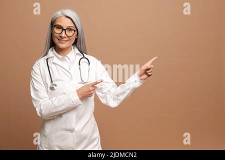 Génial, regardez ici. Portrait d'une heureuse femme d'âge moyen 50s asiatique médecin pointant de côté à un endroit vide, se tenant isolé sur fond marron, espace de copie pour publicité promotionnelle Banque D'Images
