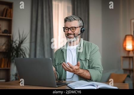 Un homme européen heureux avec une barbe dans un casque et des lunettes se gestuelle et regarde l'ordinateur portable Banque D'Images