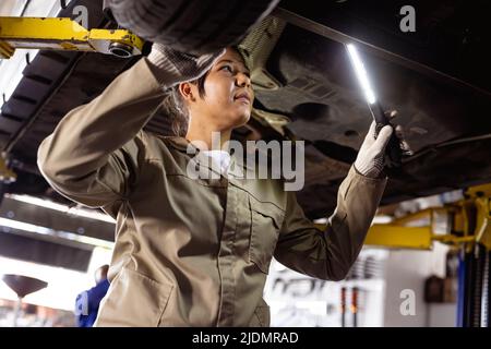 Femme asiatique moyenne utilisant un équipement d'éclairage lors de l'analyse d'une voiture défectueuse en atelier Banque D'Images