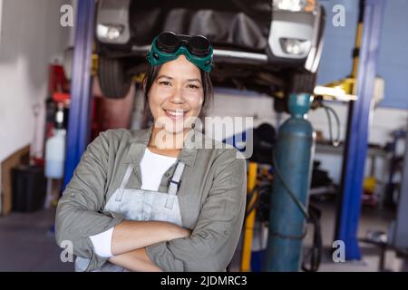 Portrait d'une soudeur asiatique moyenne adulte souriant avec bras croisés debout dans l'atelier Banque D'Images