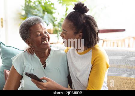 Bonne fille afro-américaine utilisant un téléphone portable tout en étant assise avec une mère âgée sur un canapé Banque D'Images