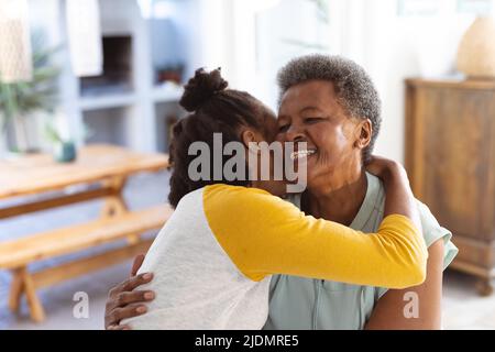 Jeune femme afro-américaine embrassant une mère âgée souriante avec des cheveux courts tout en étant assise à la maison Banque D'Images