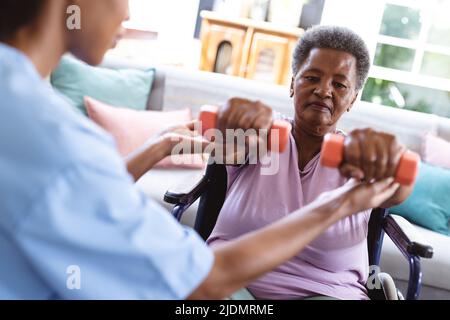 Un médecin afro-américain aide une femme âgée assise en fauteuil roulant à lever des haltères à la maison Banque D'Images