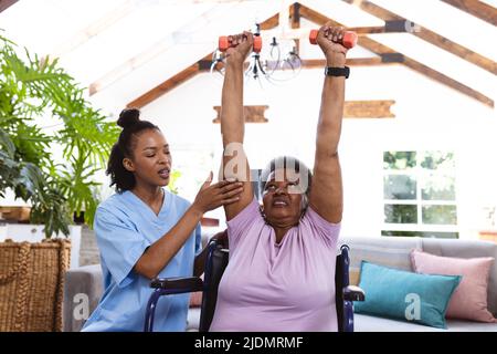Un médecin afro-américain assiste une femme âgée avec des bras levés pour soulever des haltères en fauteuil roulant Banque D'Images