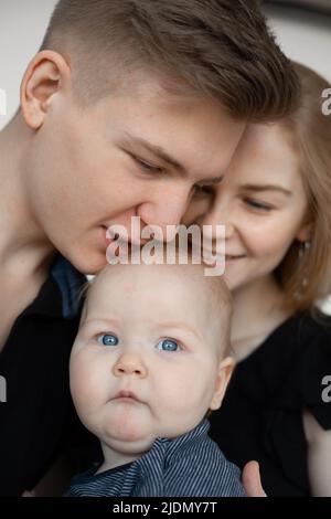 Portrait d'une jeune famille de bonne apparence dans des vêtements sombres avec un baiser de bébé chérubin sur fond blanc. Banque D'Images