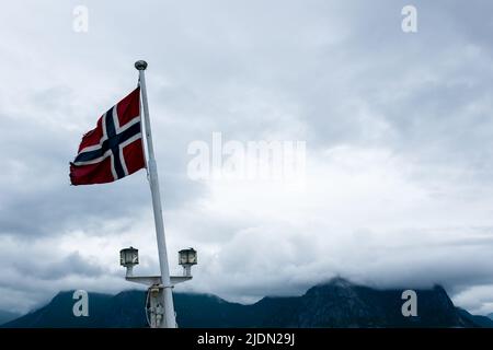 Drapeau norvégien agitant à bord d'un navire dans la mer norvégienne, océan Atlantique avec des montagnes couvertes de nuages en arrière-plan Banque D'Images