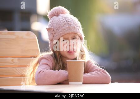 Portrait d'une petite fille mignonne dans un chapeau rose assis seul au café de rue buvant du thé de la tasse en papier Banque D'Images