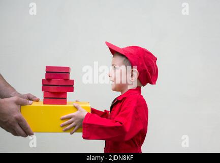 un garçon de 3-4 ans, en combinaison rouge et en casquette, récupère des boîtes aux lettres d'un adulte méconnaissable. Livraison de colis et de lettre, le petit assistant de postman, Banque D'Images