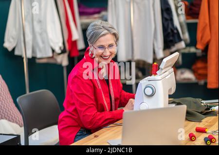 Femme souriant à l'écran d'ordinateur portable assis à la machine à coudre Banque D'Images
