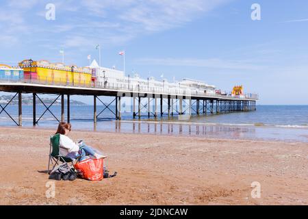 Paignton, Devon, Royaume-Uni, 14 mai 2022 : une femme seule regarde un téléphone mobile tout en étant assise dans un transat sur la plage de Paignton, près de la jetée. Banque D'Images