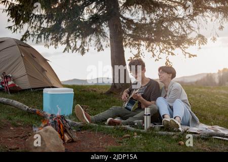 Couple joyeux de voyageurs assis au feu dans le camp et homme jouant à la guitare. Durée de vie Banque D'Images