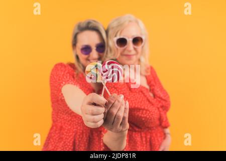 Deux femmes blonde excitées d'âge moyen portant les mêmes robes rouges pointillées et des lunettes de soleil tenant des sucettes colorées. Fond jaune, prise en studio. Photo de haute qualité Banque D'Images