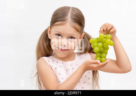 Belle petite fille caucasienne de 6 ans tenant un bouquet de raisins de table verts sur fond blanc regardant l'appareil photo Banque D'Images