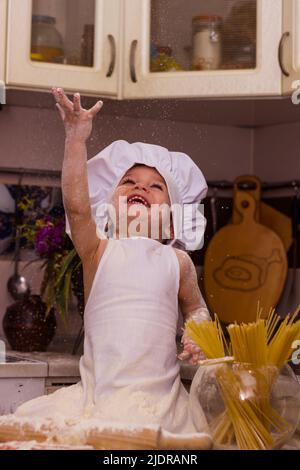 Un enfant en costume de cuisinier est assis dans la cuisine sur une table et sourit Banque D'Images