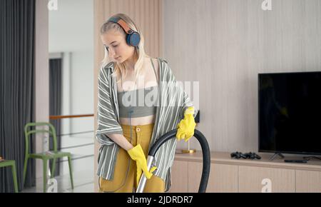 Une jeune fille se moque de nettoyer le sol avec un aspirateur. Bonne femme qui fait du ménage à la maison Profitez de la musique avec des écouteurs. Banque D'Images